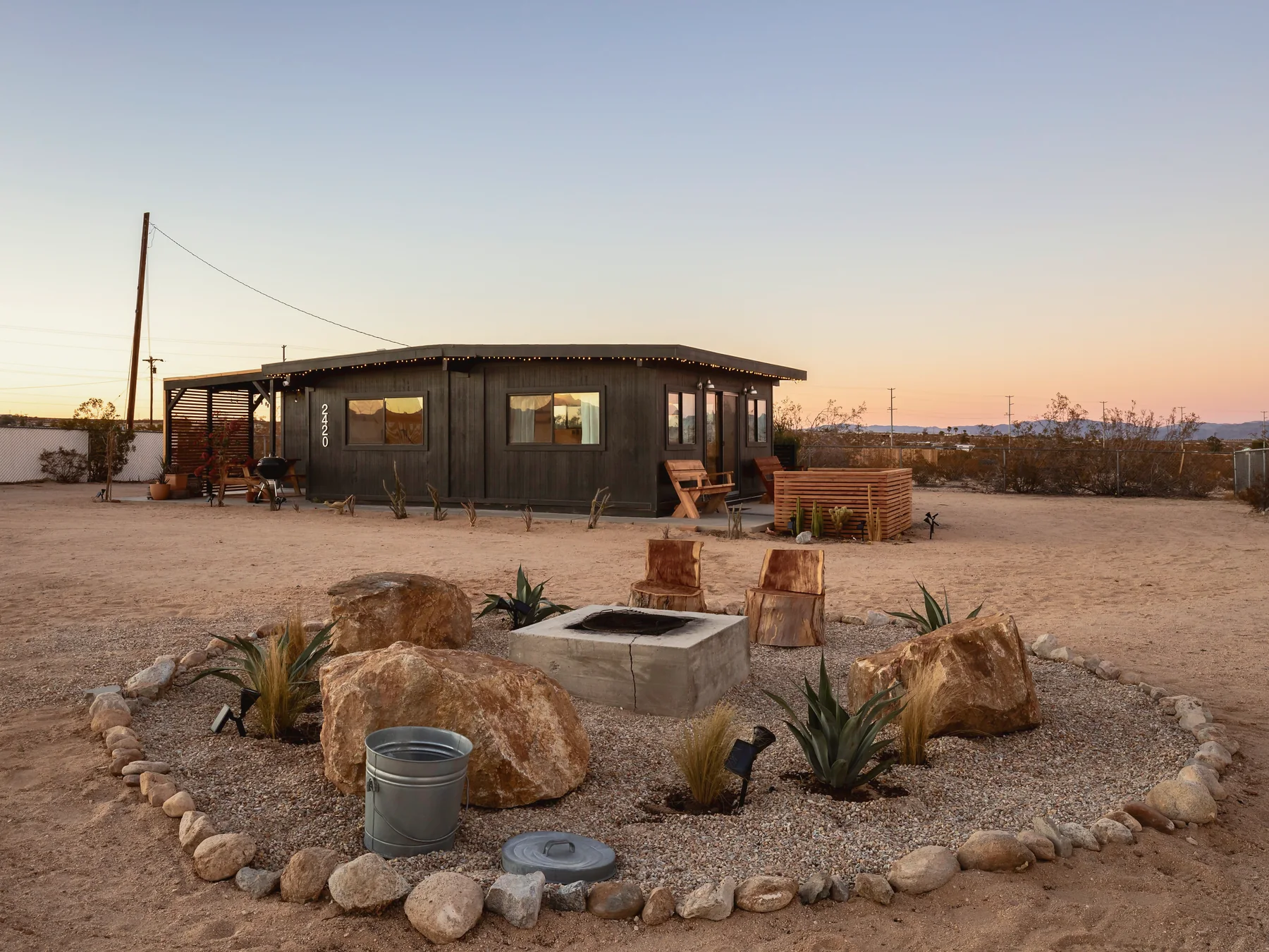 Stone-ringed fire pit with boulders and agaves, the black cabin behind it at dusk